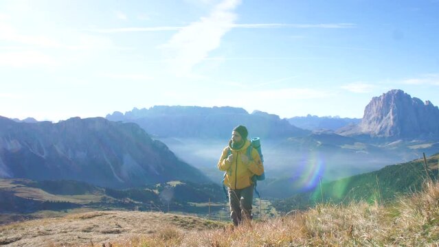 Young man hiking at Seceda mountain arriving to peak at sunrise. Backpack, yellow jacket, boots, beanie. Traveling to puez Odle, Dolomites, Trentino, Italy.