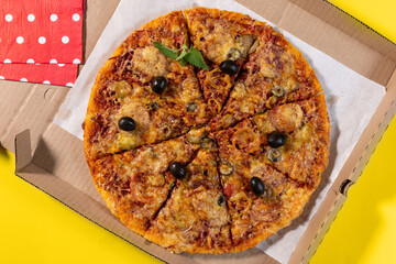 Pizza with vegetables and sausage in a cardboard box for home delivery on a yellow background