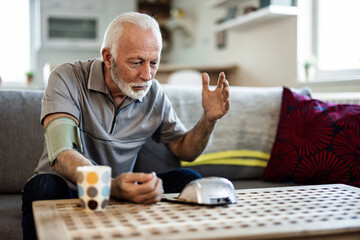 Senior man sitting in his living room on a sofa and checking his blood pressure. Photo of mature man measuring her blood pressure at home.