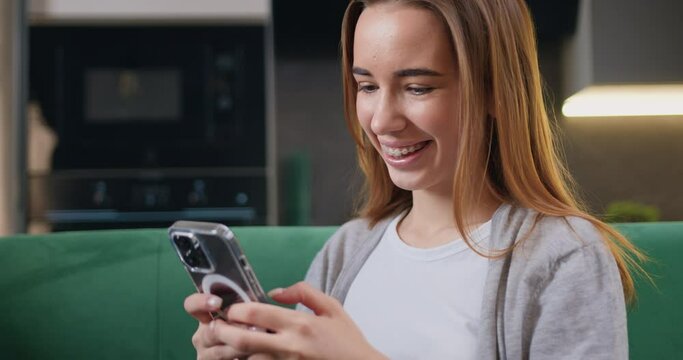 Cheerful young woman sitting on a sofa and using smartphone in modern room. Beautiful girl smiling and thinking while chatting on mobile phone.