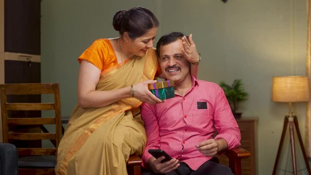 excited wife giving gift to husband by closing eyes while sitting on chair at home - concept of togetherness, happt long relationship and wedding anniversary.