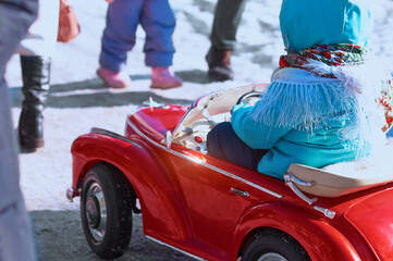 Girl driving a red children's car cabriolet among pedestrians. Baby girl is dressed in warm clothes...
