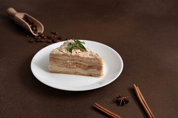 Delicious Napoleon Cake with cream on a white plate decorated with a sprig of mint on a Brown Background. A cinnamon stick, badyan, coffee beans on a brown background. Copy space