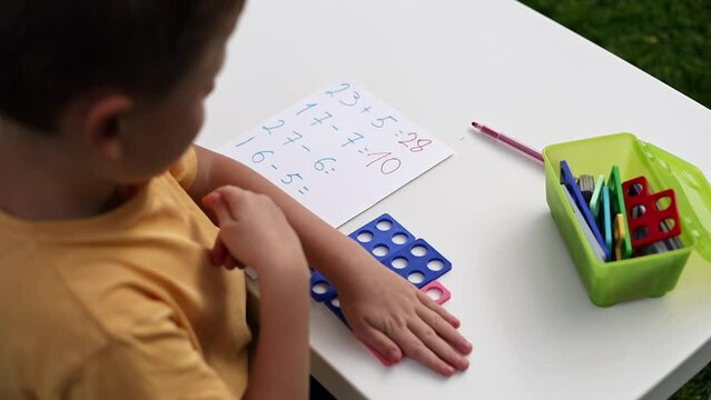 Child with autism has math therapy lesson: uses colorful Numicon shapes, counts, writes answers on paper; sits at white table in sunny garden with bright green grass
