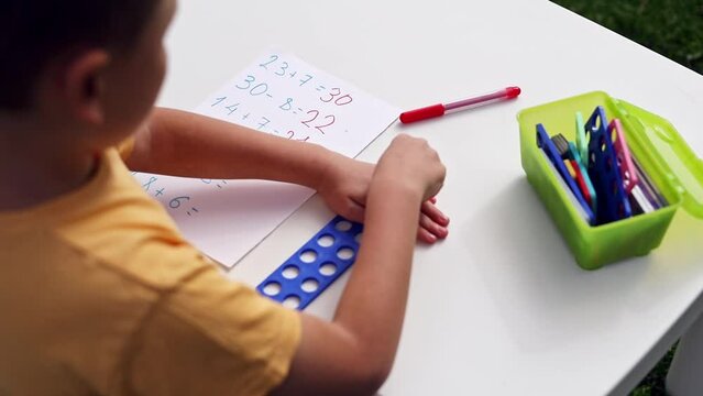 Child with autism has math therapy lesson: uses colorful Numicon shapes, counts, writes answers on paper; sits at white table in sunny garden with bright green grass