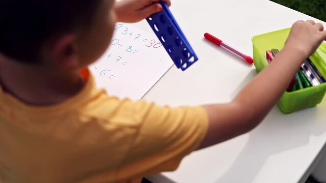 Child with autism has math therapy lesson: uses colorful Numicon shapes, counts, writes answers on paper; sits at white table in sunny garden with bright green grass
