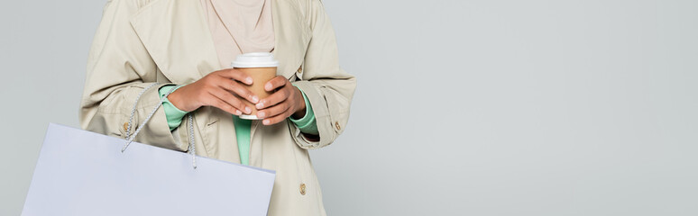 partial view of multiracial woman in trench coat holding shopping bag and disposable cup isolated on grey, banner.