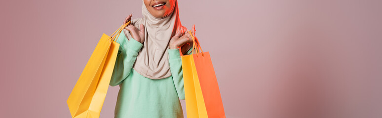 cropped view of happy multiracial muslim woman holding yellow shopping bags on pinkish grey...