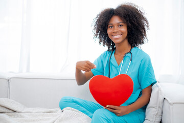 Portrait of African American doctor holding red heart pillow pointing at it with smile of kindness,...