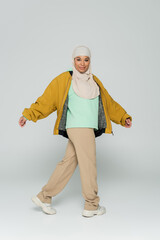 full length of cheerful multiracial muslim woman in yellow bomber jacket and beige pants walking on grey background.