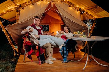 Happy funny couple relaxing in glamping on summer evening and drinking wine near cozy bonfire. Luxury camping tent for outdoor recreation and recreation. Lifestyle concept
