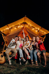 Obraz na płótnie Canvas Happy group of friends with Welsh Corgi Pembroke dogs relaxing in glamping on summer evening near cozy bonfire. Luxury camping tent for outdoor recreation and recreation. Lifestyle concept