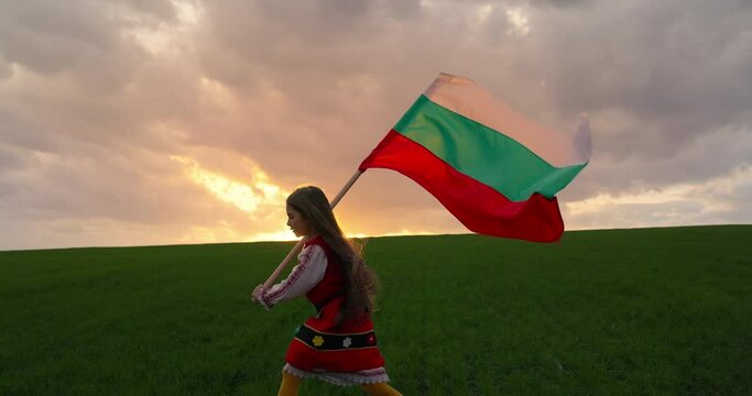Beautiful bulgarian girl in folklore ethnic costume and bulgarian flag running outdoor on green agriculture field. Freedom, beauty and bulgarian independence and pride concept, 4k video