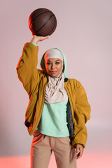stylish multiracial woman in hijab and yellow bomber jacket standing with basketball in raised hand...