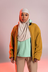 young multiracial muslim woman in yellow bomber jacket and hijab looking at camera on grey and pink...