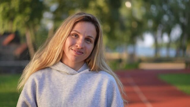 Smiling sportswoman touching hair looking at camera smiling standing on sports track in sunshine. Portrait of fit Caucasian woman posing at sunset in park after workout training