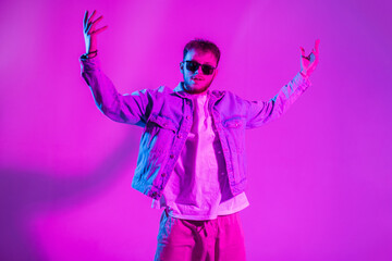 Creative handsome fashion dancer man with sunglasses in stylish denim clothes dance in colorful studio with pink and neon light