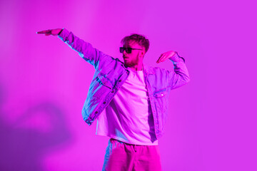 Handsome stylish fashion dancer man with sunglasses in a denim jacket dancing in a creative colorful studio with pink and neon lights