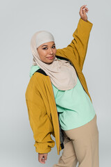 fashionable multiracial woman in yellow bomber jacket and hijab posing with raised hand isolated on grey.