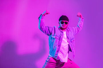 Stylish handsome professional hip hop dancer man with cap and sunglasses in trendy fashion denim clothes dancing in studio with creative pink and neon light