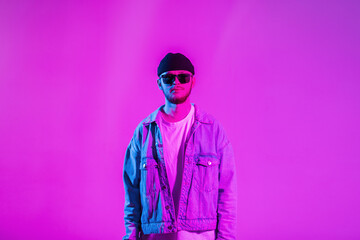 Stylish handsome creative young guy with a cap and sunglasses in a trendy fashion denim jacket in a studio with pink and neon lights