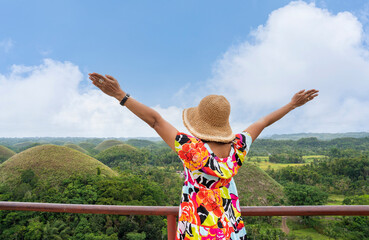 Female tourist in tourist hat walking up hill raising hands feeling free, happy, vacation, chocolate hills landmark of philippines.