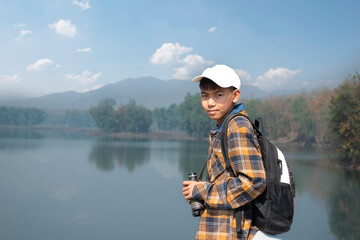 Asian boy in plaid shirt wears cap and has a backpack, holding a binoculars, standing on ridge reservoir in local national park to observe birds on tree branches and on sky and to watch fish in lake.