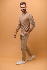 Fototapeta na wymiar Full length portrait of handsome bearded man in beige pullover is holding hand in pockets and walking, guy in stylish eyeglasses looking at the camera with pleasant smile, isolated studio shot