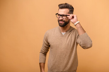 Attractive man isolated on beige background, guy standing and looks aside with friendly smile, male entrepreneur in casual wear adjusting stylish eyeglasses