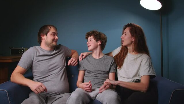 Caring parents sit on a blue sofa with their teenage son and communicate on various topics, carefully hugging and kissing their child. Good parents and the transitional age of a teenager of 14 years