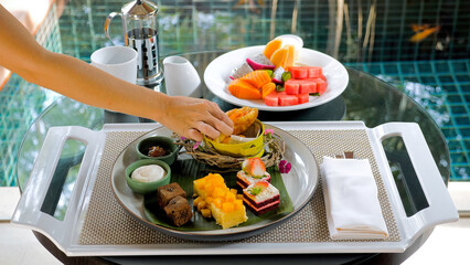 Luxurious breakfast on villa terrace with tropical fruits, desserts, and cakes. Woman eating a fresh croissant with smoked salmon. Perfect start to a vacation day. Beautiful served.