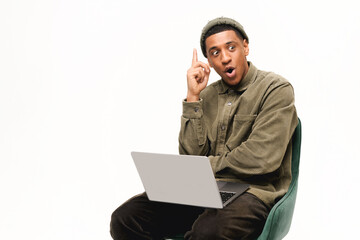 Excited young african-american man in trend hat and shirt came up with new idea or solution, pointing finger up, sitting with laptop on laps isolated over white background