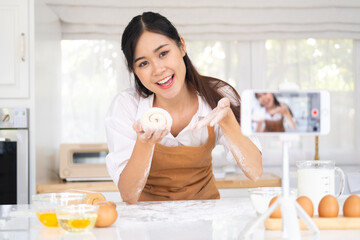 Portrait of smiling young woman food vlogger showing process of baking in live stream, standing in front of smart phone on tripod, broadcasting. Indoor studio shot on kitchen background.