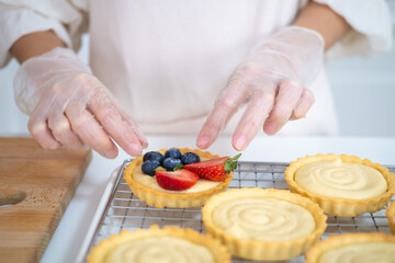 Closeup of hands in cooking gloves decorating freshly baked tart with blueberries strawberry fresh fruit.housewife baker wear apron making fruit tart. homemade bakery at home.