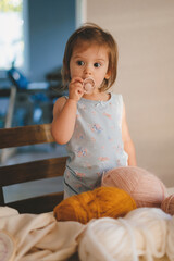 A cute baby girl standing on a chair near table full of tangles of knitting threads. Hendmeid of...