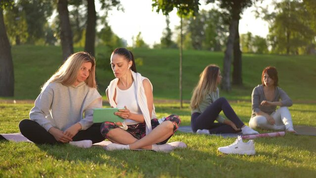 Positive fit Caucasian women with digital tablet sitting on green lawn at sunset talking in slow motion. Blurred friends chatting at background in sunshine. Sport and friendship concept