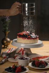 Chef's hand adding powdered sugar on a delicious cake with berries on a white ceramic stand