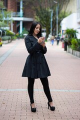 Vertical shot of a young beautiful female in a black jacket and heels using her phone