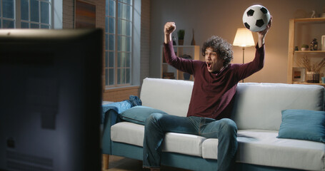 Expressive Spain man football team supporter emotionally watching a soccer game. Hipster guy with curly hair watching tv at home alone 