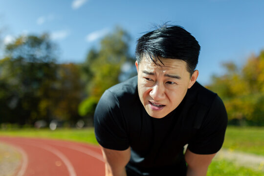 Asian sportsman tired after running and active physical exercise, man taking breath and rest on sunny day in stadium, overtired after running. - Powered by Adobe