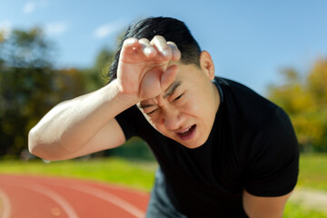 Naklejka premium Close-up of sportsman having severe headache and dizziness, asian man in stadium doing fitness and active exercise, male runner in sportswear overworked by training on sunny day.