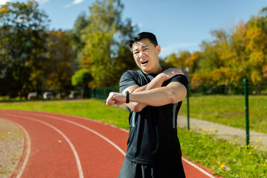 Asian sportsman stretches his shoulder, man in the stadium after running and active physical exercises stretches his arm joints, has shoulder pain, arm muscle spasm.