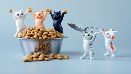 Cute toy kittens with raised paws carry a spoon to a metal bowl of dry pet food. Concept of the...