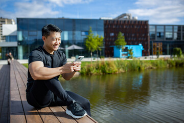 Asian sportsman resting after training sitting on a bench in the park near the lake and using the...