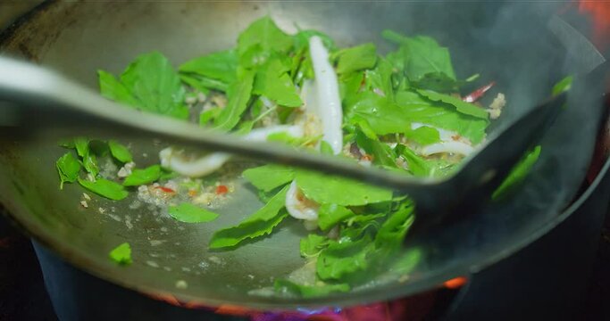 Close up cooking stir fried minced pork and squid with green Thai Basil Leaves chili peppers and garlic in a frying pan wok on hot fire flame from gas in the kitchen. Delicious Thai food.