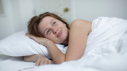 Young happy beautiful sleepy woman in the bed in bedroom at home in the morning lying under white blanket, enjoy resting, sleep well, wake up and smile. Good morning