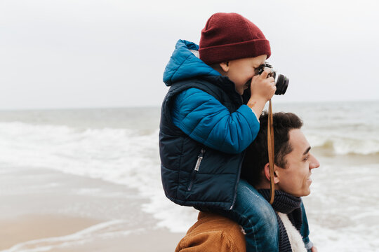 Boy sitting on his father's shoulders and taking pictures while spending time together on beach