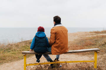 Back view of father and son enjoying sea view while sitting on bench at beach
