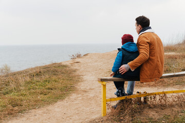 Back view of father and son enjoying sea view while sitting on bench at beach