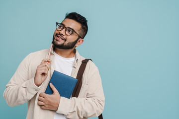Pensive indian man looking away while standing with pencil and notebook isolated over blue wall